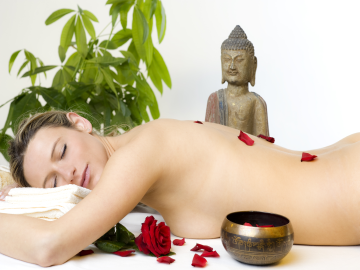 Eine auf dem Bauch liegende Frau mit nacktem Oberkörper auf einer Wellnessliege in einem hellen Raum mit Rosenblättern auf dem Rücken und Buddhastatue im Hintergrund